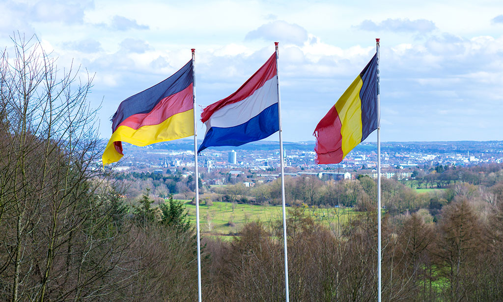 Die deutsche, luxemburgische und belgische Fahne wehen auf langen Masten im Wind, im Hintergrund sieht man Wald und eine Ortschaft. 