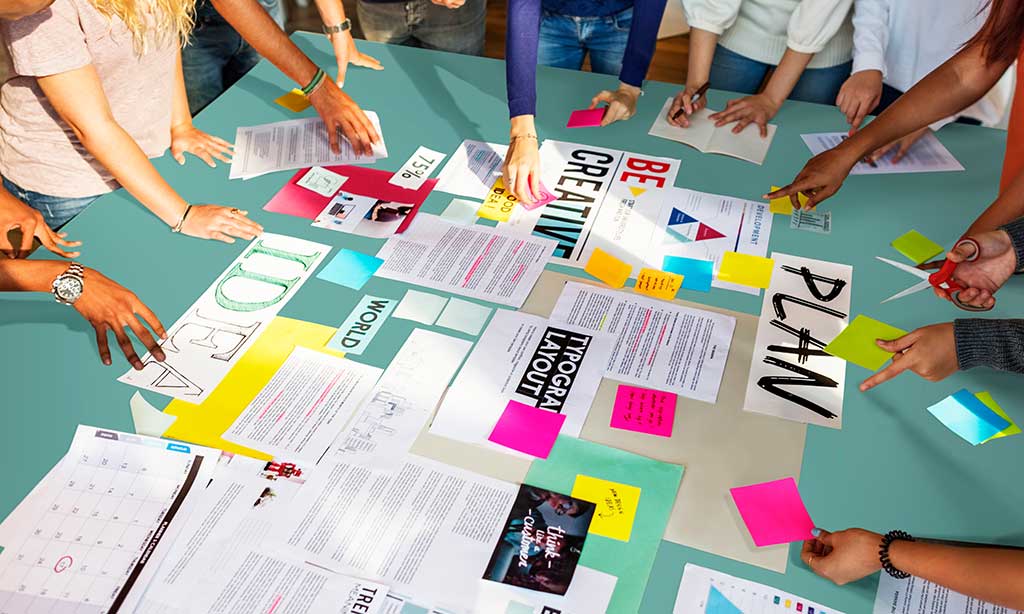 Mehrere Personen stehen um einen Tisch auf dem bunte Klebezettel und Papiere liegen und brainstormen. 