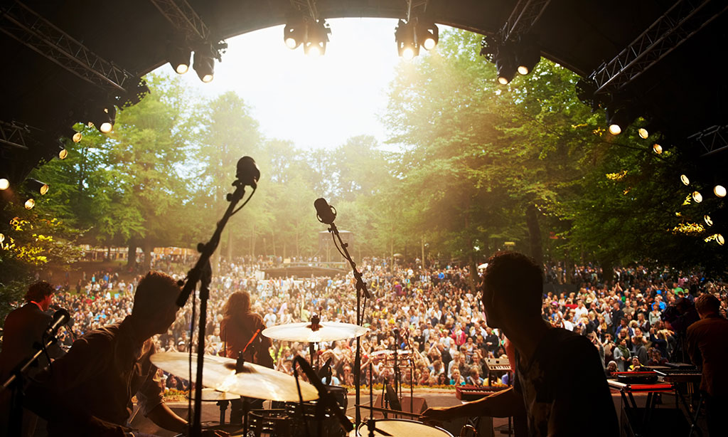 Eine Open-Air-Bühne mit Musikern und eine Menschenmenge in einem grünen, sonnenbeschienenen Park. 